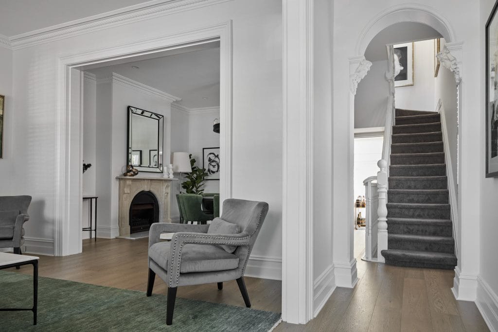 A modern living room with gray armchairs, fireplace, mirror, and a carpeted staircase beside white walls; Emma Blomfield sits nearby.
