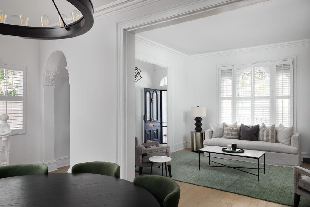 A modern living room with white walls, gray sofa, green rug, black table; dining area with green chairs; sunlight streams in.