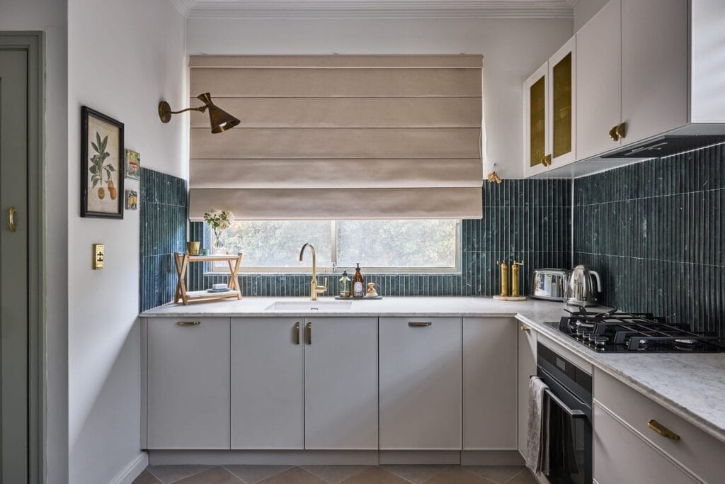 Emma Blomfield stands in her modern kitchen featuring white cabinets, a dark green tiled backsplash, brass fixtures, and a roman shade covering the window above the sink, with a painting and shelf of dishes on the left wall that showcase tasteful brand collaborations.