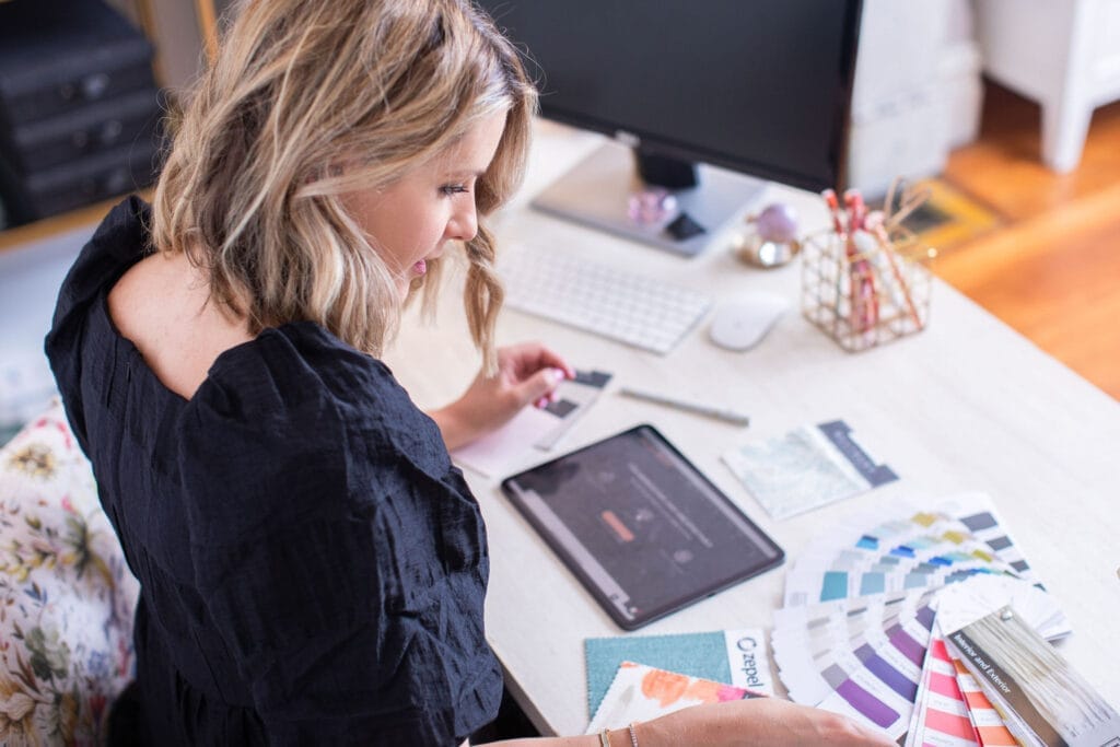 Emma Blomfield, with her blonde hair and black top, sits at a desk filled with color swatches, paper samples, an iPad, and office supplies, clearly immersed in her interior decorating business.