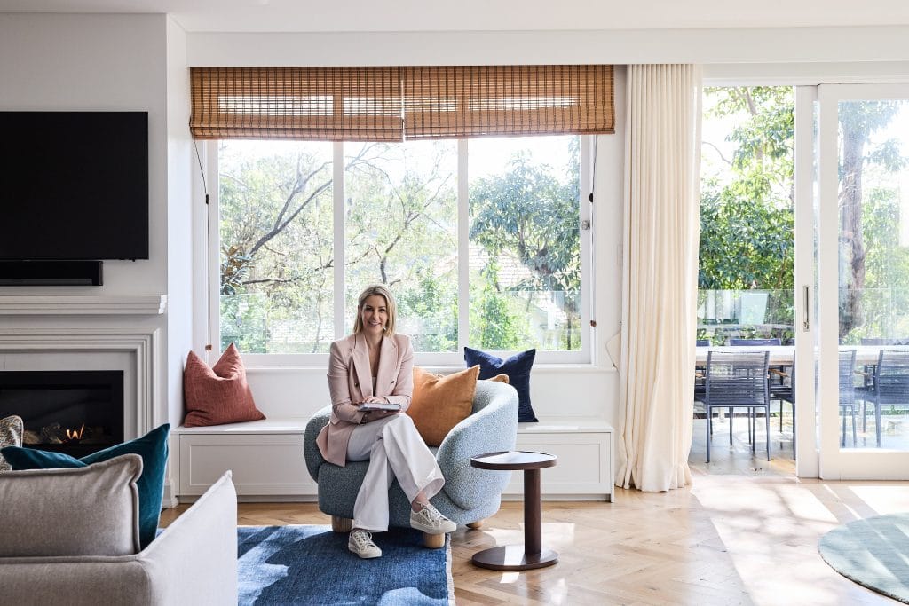 Emma Blomfield sits on a blue chair in a modern living room with large windows and a view of the patio.