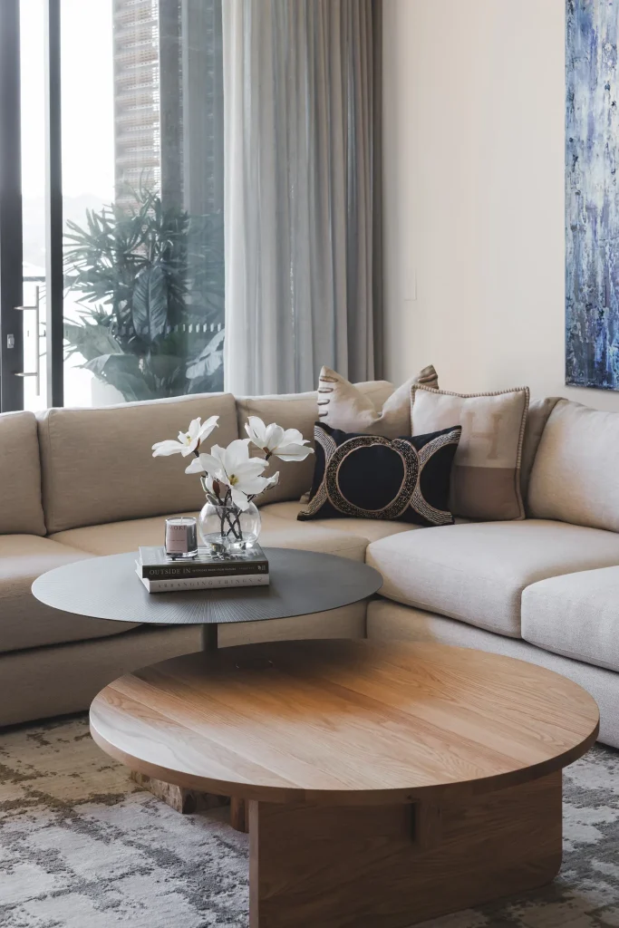 A modern living room with a beige sectional, round tables, white flowers, cushions, and sheer curtains near the window.
