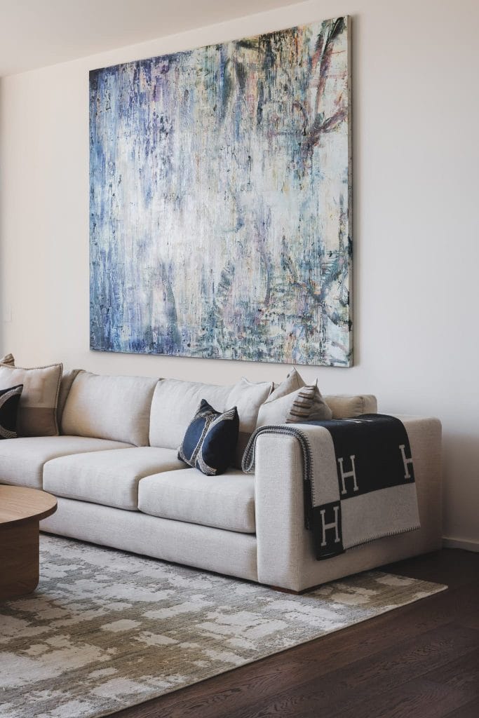 A beige sofa with dark pillows and a blanket sits on a patterned rug beneath an abstract painting in this modern living room.