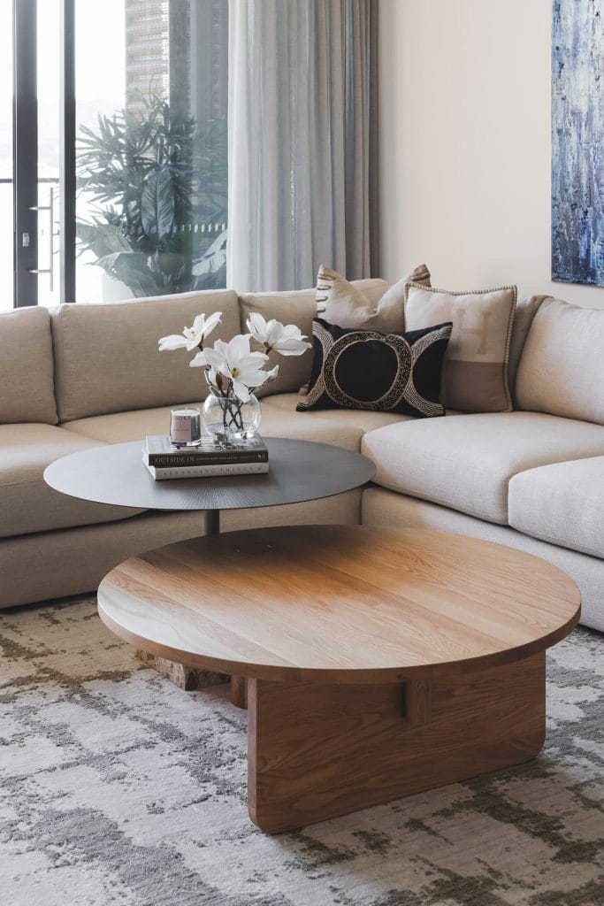 Modern living room with beige sectional, round coffee tables, white flowers, and sheer-curtained windows styled by an expert.