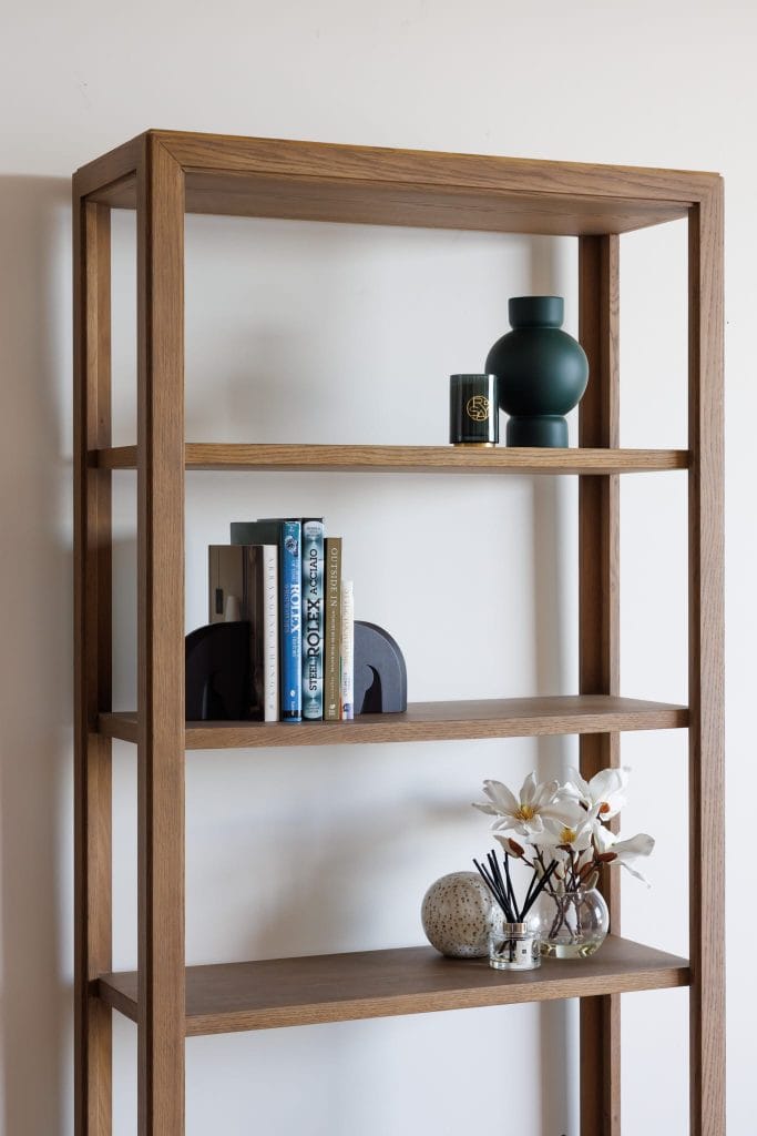 A wooden bookshelf with books, a vase, candle, decorative orb, and flowers decorates a room with a plain white wall.