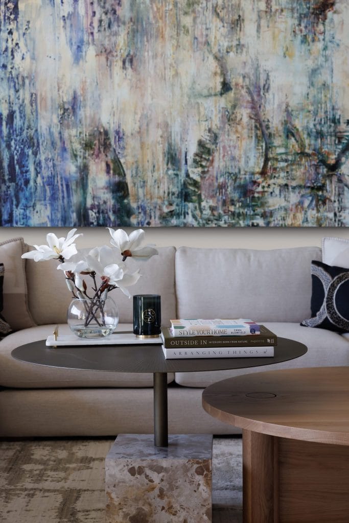 A modern living room with a beige sofa, two round tables, white flowers in a glass vase, books, and abstract wall art.