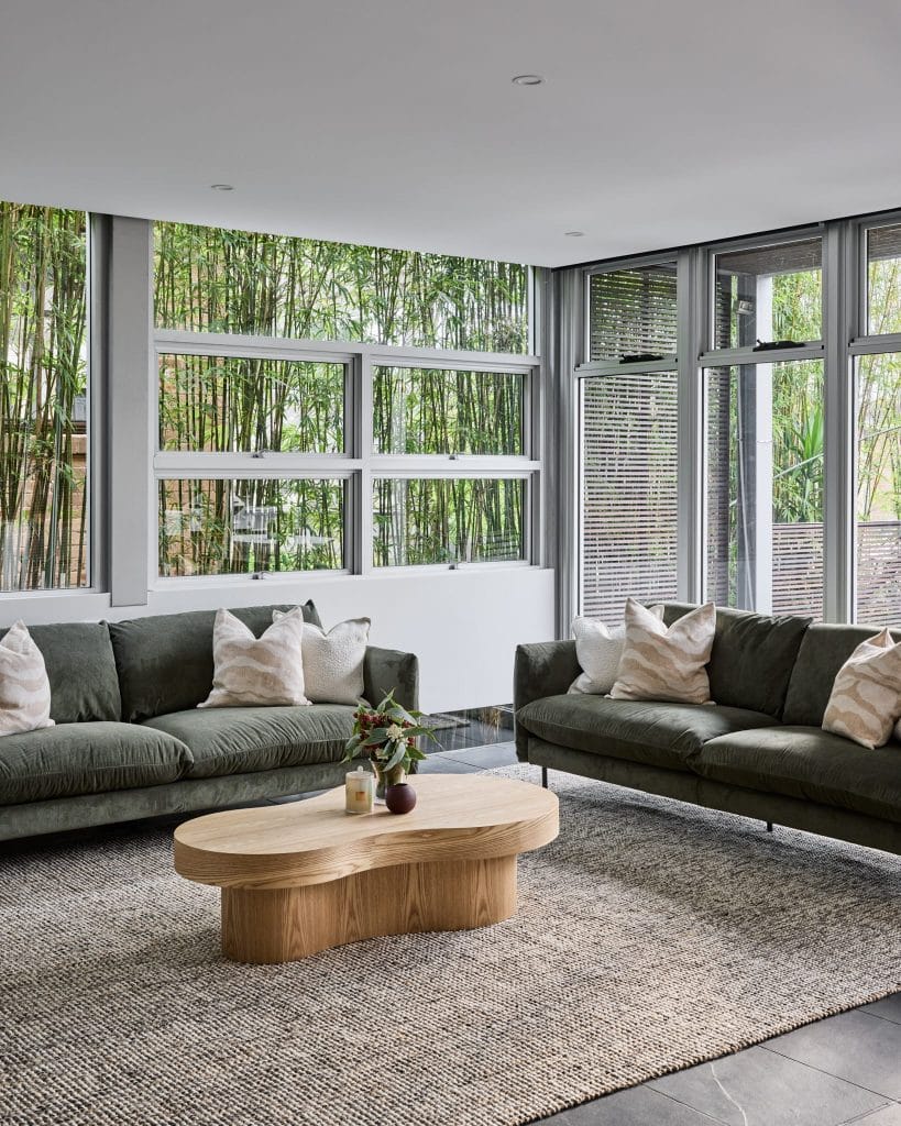 Modern living room with two dark green sofas, a light wood coffee table, and large windows overlooking bamboo plants.