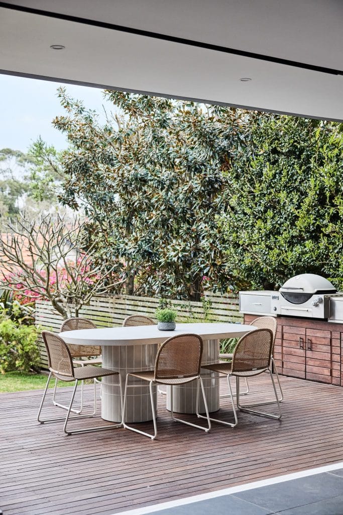 Outdoor patio with white table, six wicker chairs, barbecue grill, and lush greenery, styled effortlessly by Emma Blomfield.