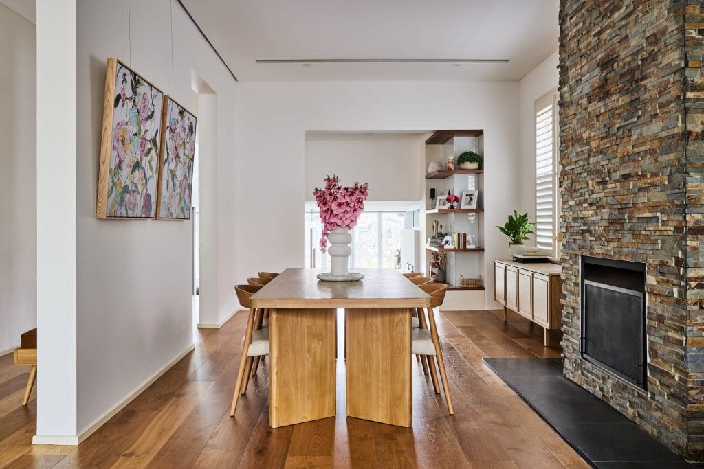 Modern dining room with wood table, six chairs, pink flowers, stone fireplace, wall art, wood floors, and built-in shelves.