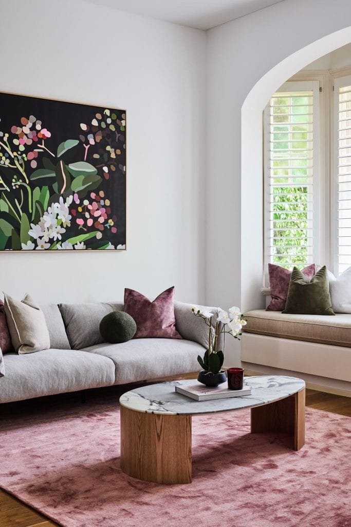 A modern living room with a gray sofa, colorful cushions, marble table, pink rug, floral art, and window; Emma Blomfield is present.