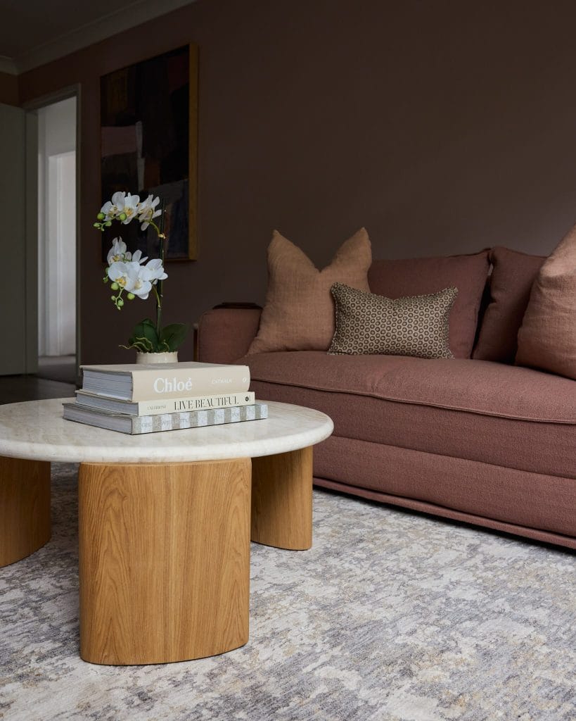 A modern living room with a mauve sofa, patterned pillows, and a round coffee table adorned with books and a white orchid.