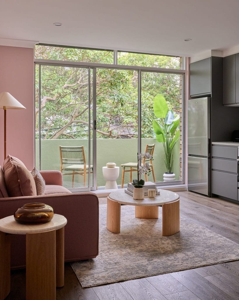 A modern living room with a sofa and tables opens to a balcony with two chairs and lush trees through large sliding glass doors.