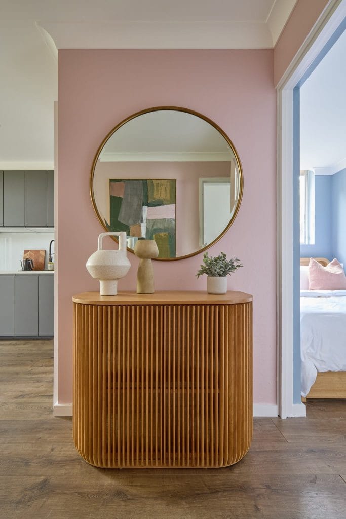 A wooden console with decor and a potted plant sits below a round mirror on a pink wall, hinting at kitchen and bedroom beyond.