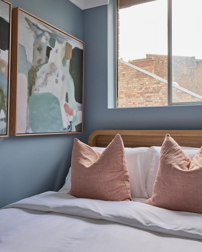 A neatly made bed with pink pillows, white bedding, abstract art on a blue wall, and a window revealing brick buildings outside.