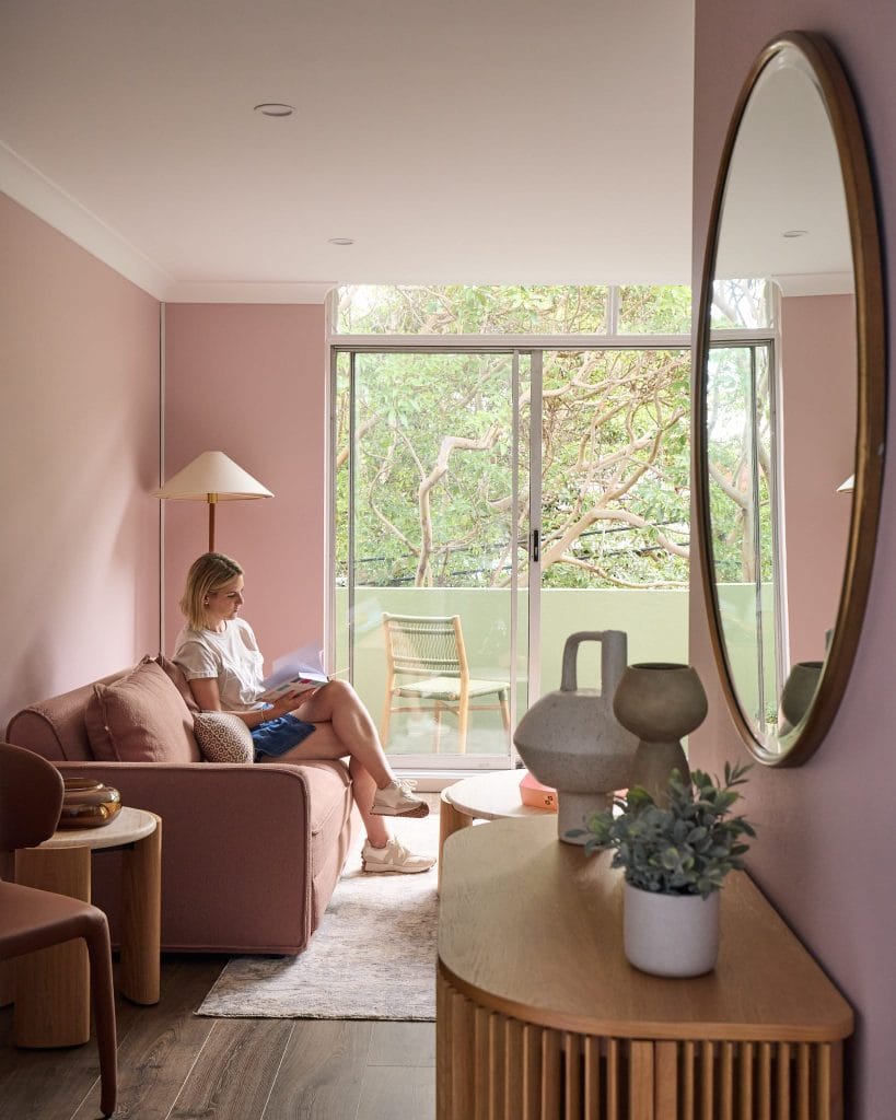 A woman sits on a sofa reading in a chic pink living room with a large round mirror and glass doors to a balcony with greenery.