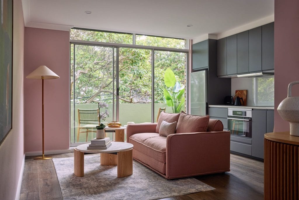Modern living room with pink sofa, round tables, floor lamp, large windows with greenery, and compact kitchen on the right.