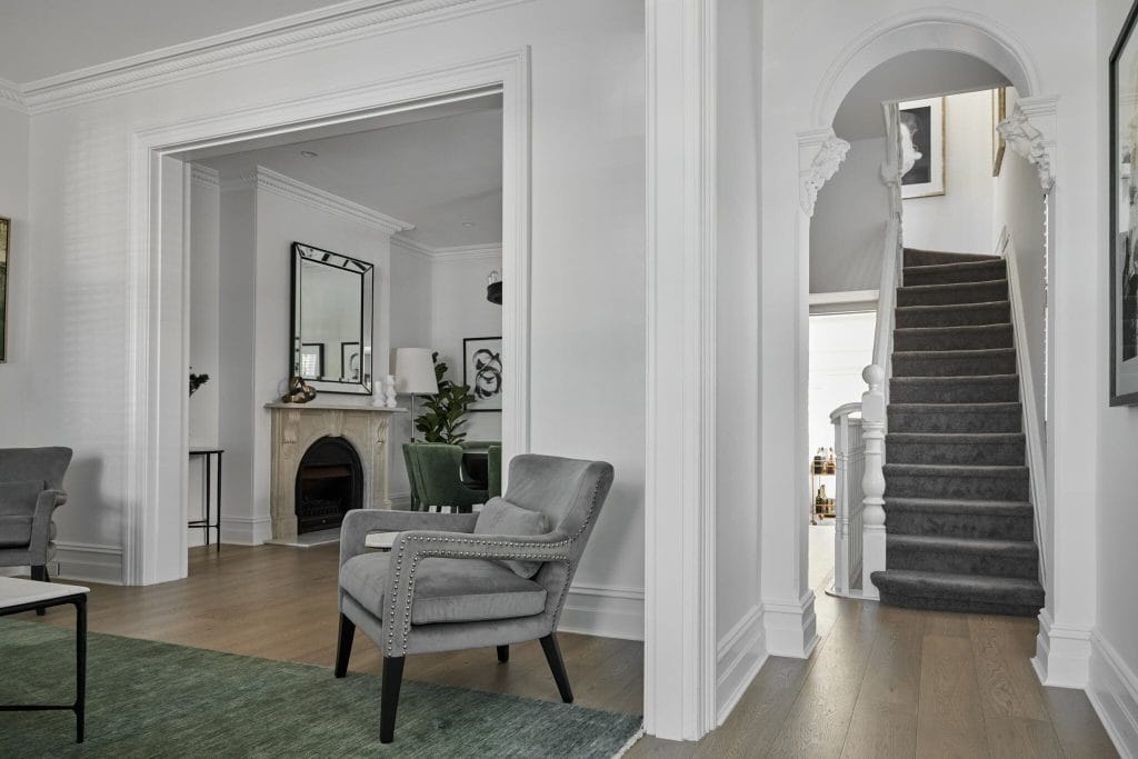 A stylish living room features grey armchairs, a fireplace, grey-carpeted stairs, and white walls with molding.
