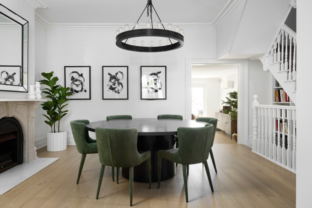 Modern dining room featuring a round black table, six green velvet chairs, abstract art, and ring chandelier on wood flooring.