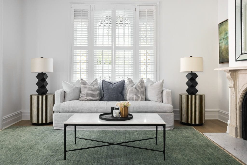 A stylish living room with a gray sofa, green rug, coffee table, and black lamps by a window; Emma Blomfield sits inside.