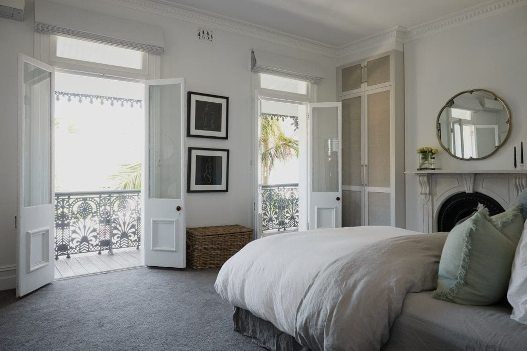 Bright bedroom with balcony, double doors, neutral bedding, fireplace, round mirror, framed art; styled for inspiration.
