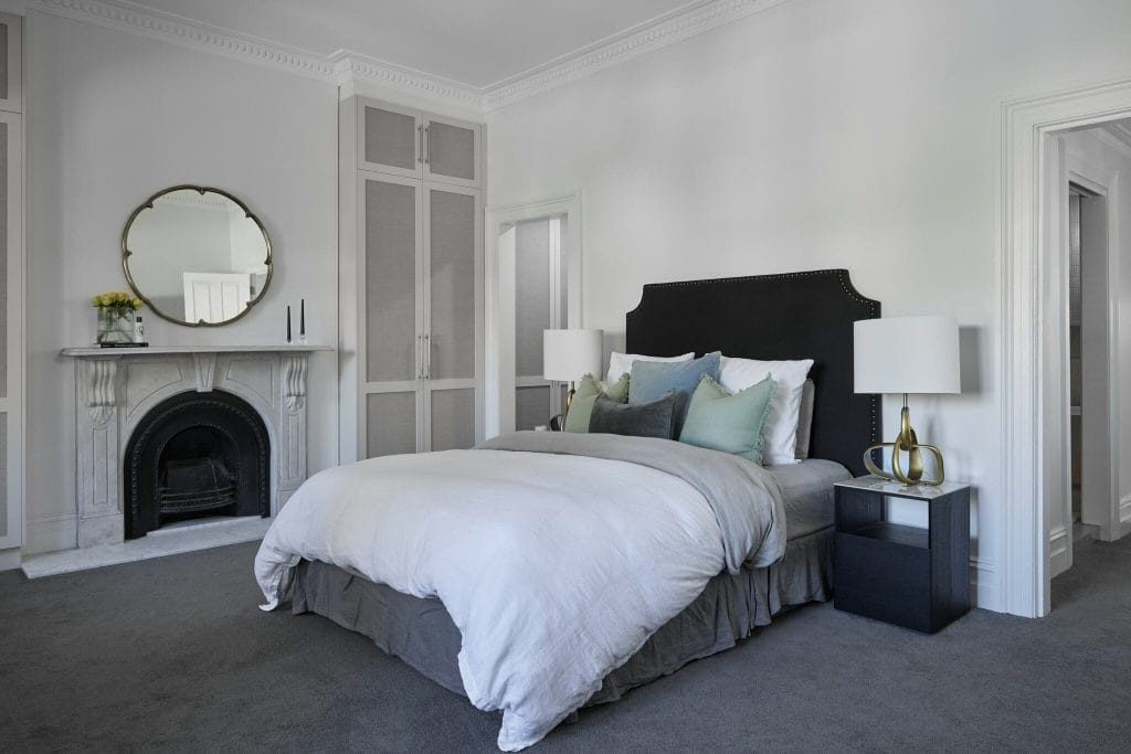 Modern bedroom with black upholstered bed, white bedding, bedside tables, lamps, fireplace, and a round mirror over the mantel.