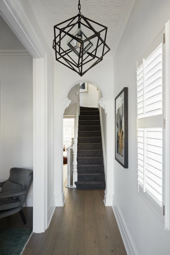 A narrow hallway with white walls, a geometric pendant light, an arched doorway, and a carpeted staircase shows modern elegance.