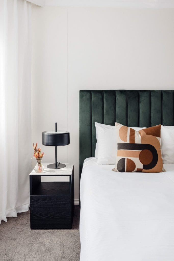 A modern bedroom with a green headboard, geometric pillow, white bedding, black table, lamp, and vase; Emma Blomfield present.