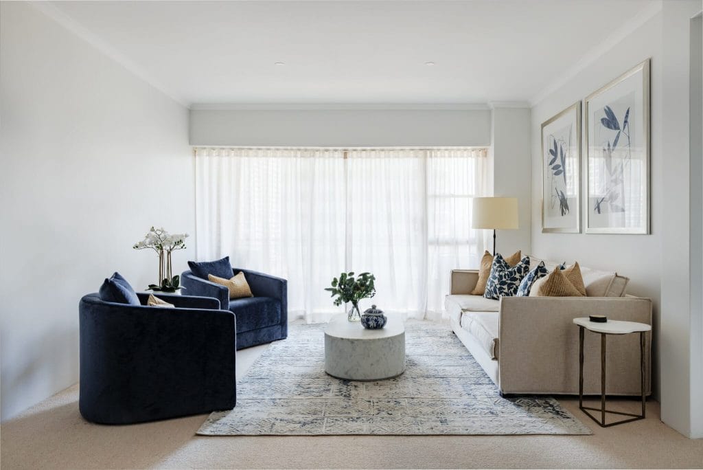 A modern living room with a beige sofa, navy armchairs, marble coffee table, patterned rug, and sunlit sheer curtains; Emma Blomfield is present if pictured.