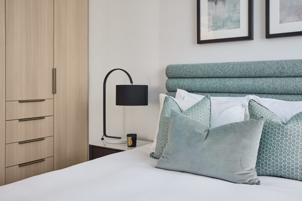 Modern bedroom in Cremorne featuring a teal upholstered headboard, white bedding, a black lamp on the nightstand, and a wooden wardrobe with drawers, accented by two framed artworks on the wall.