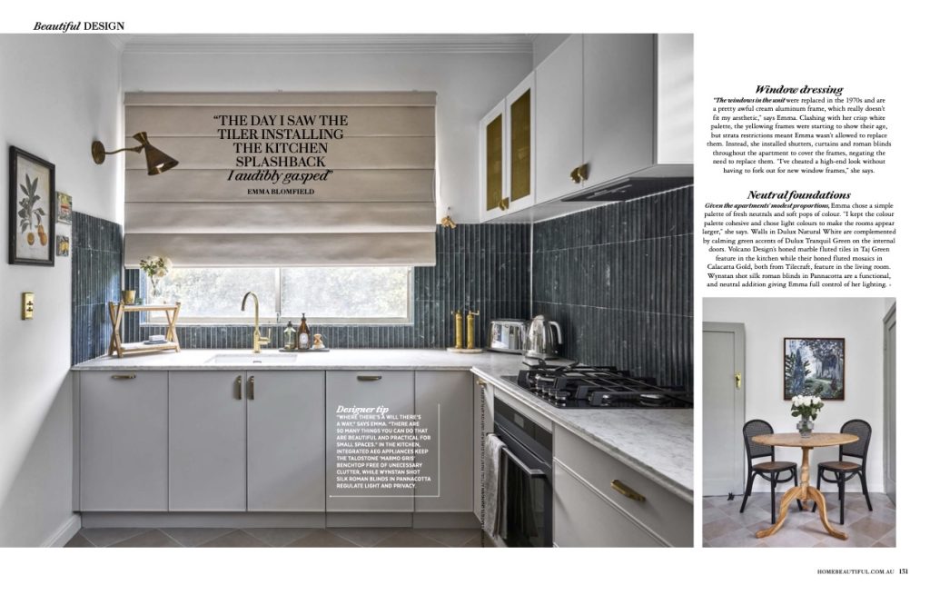 Modern kitchen with white cabinets, dark green tile backsplash, gold fixtures, and a beige Roman shade with a printed quote, naturally lit by a window above the sink.