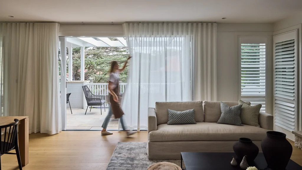 Emma Blomfield pulls sheer curtains open in a modern living room with a sofa, cushions, and large windows leading to an outdoor patio.