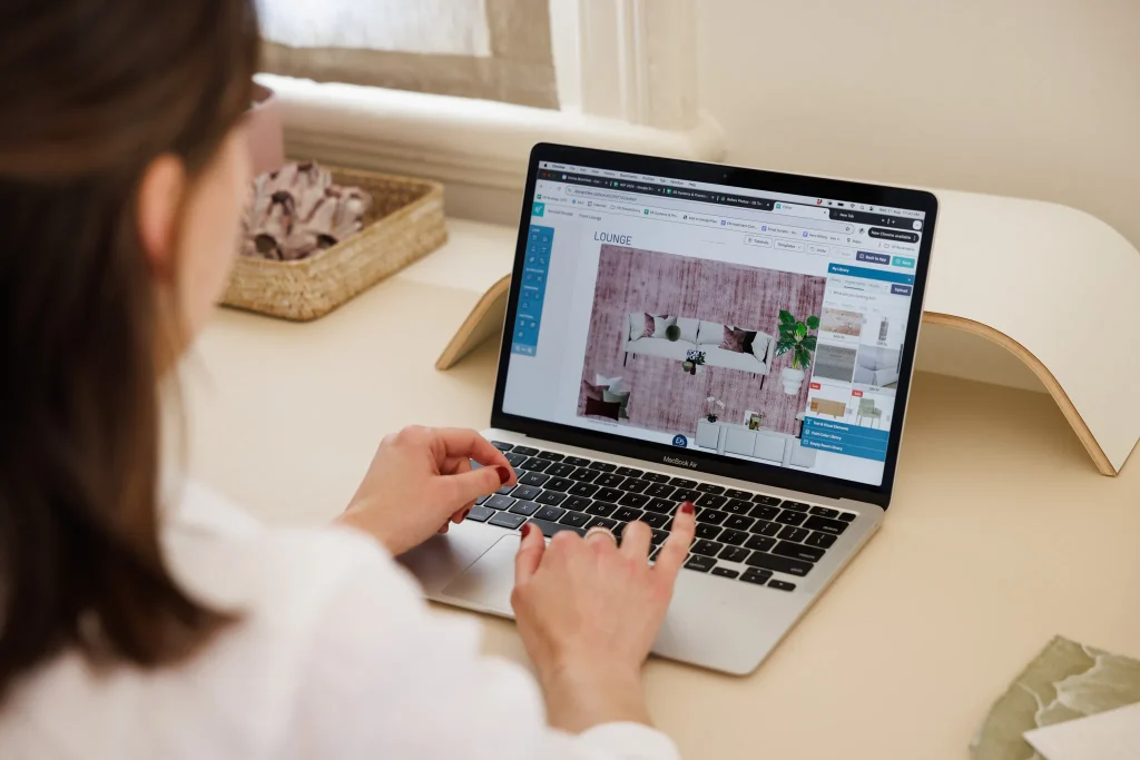 Emma Blomfield, a blonde woman, uses a laptop to browse an interior design site showing a living room with a pink rug, white sofa, and green plants.