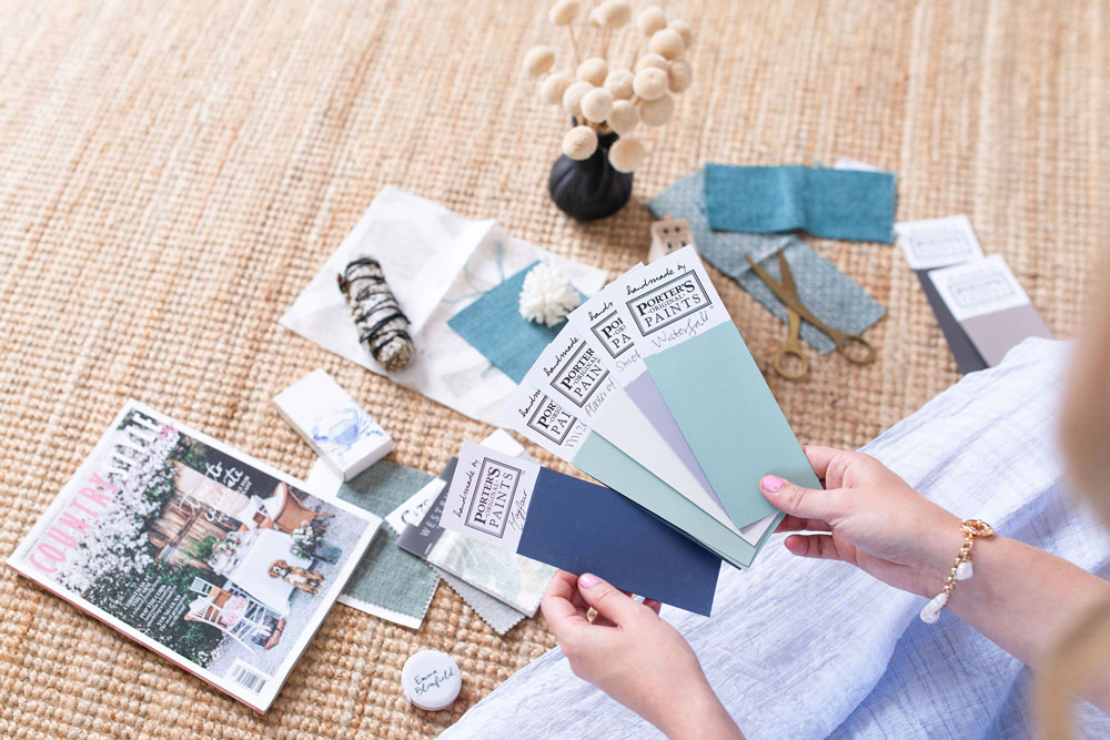 Emma Blomfield holds paint swatches over a woven rug, surrounded by fabric samples, magazines, scissors, and decor.