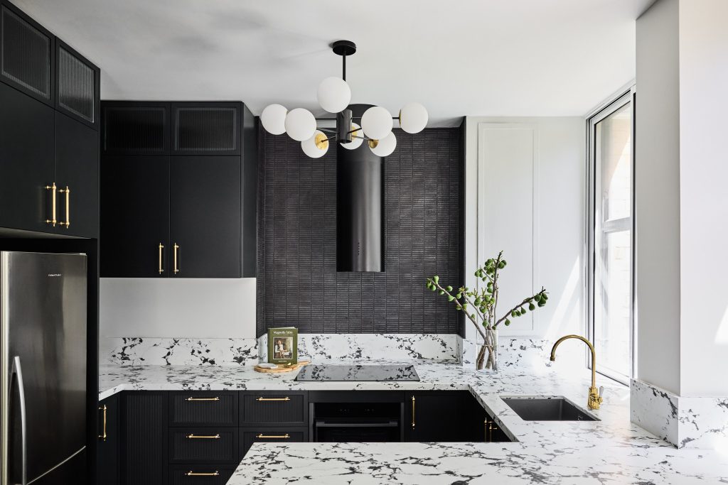 Modern kitchen with black cabinets, white and black marble countertops, gold hardware, a white globe light fixture, and a vase of green branches by the window; if present, Emma Blomfield stands nearby.