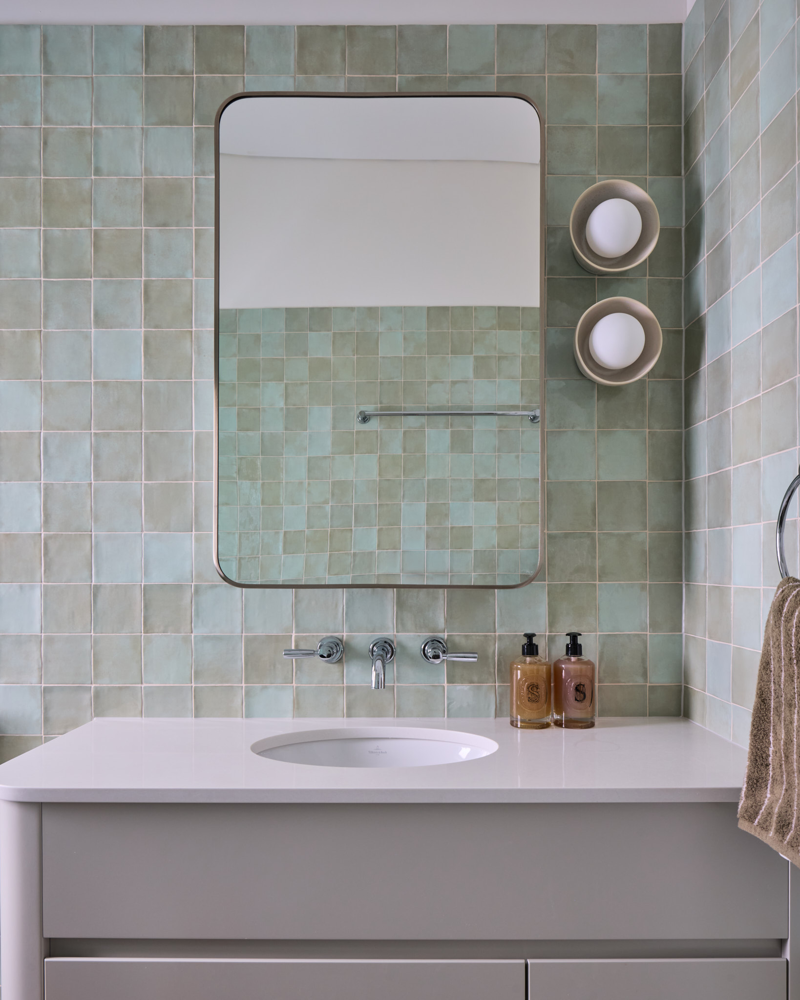 Modern Mosman bathroom featuring a light green tiled wall, rectangular mirror, wall-mounted faucet, white sink, soap bottles, towel and a round shelf with two rolled towels.