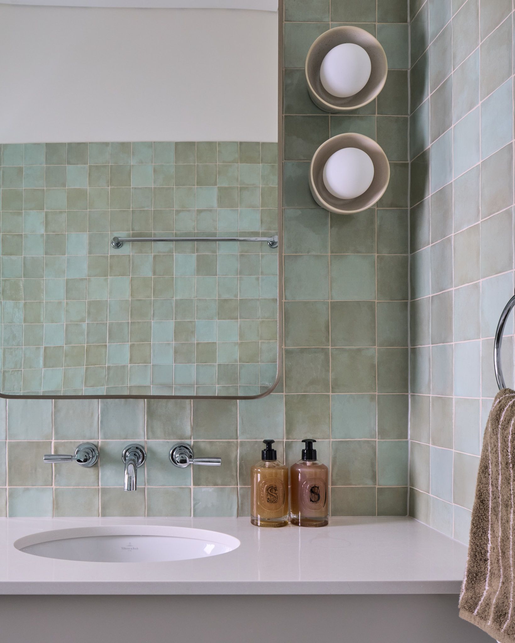 Modern bathroom sink with green tiles, two soap dispensers, a towel on a ring, round shelves with white items, and a mirror above the counter.