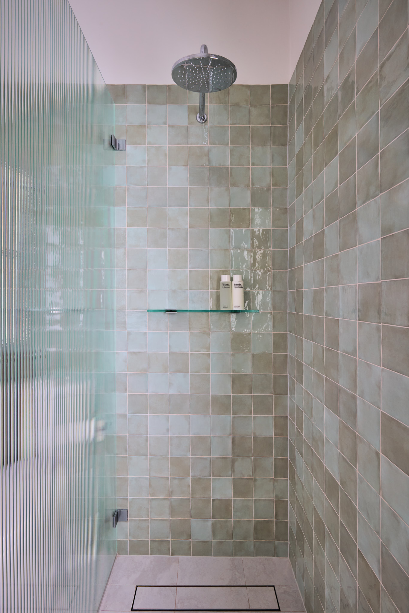 Modern shower with greenish square tiles, glass shelf of bottles, rainfall showerhead, and ribbed glass partition on the left.