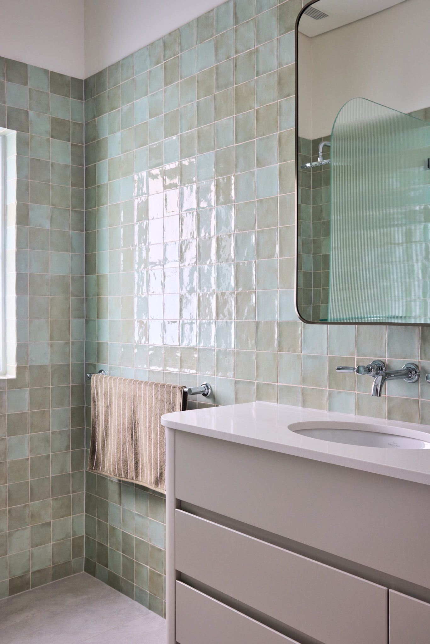Bathroom with pale green square tiles, a rounded rectangular mirror, white vanity, and towel on the wall exudes calm sophistication in this refreshed Mosman Home.