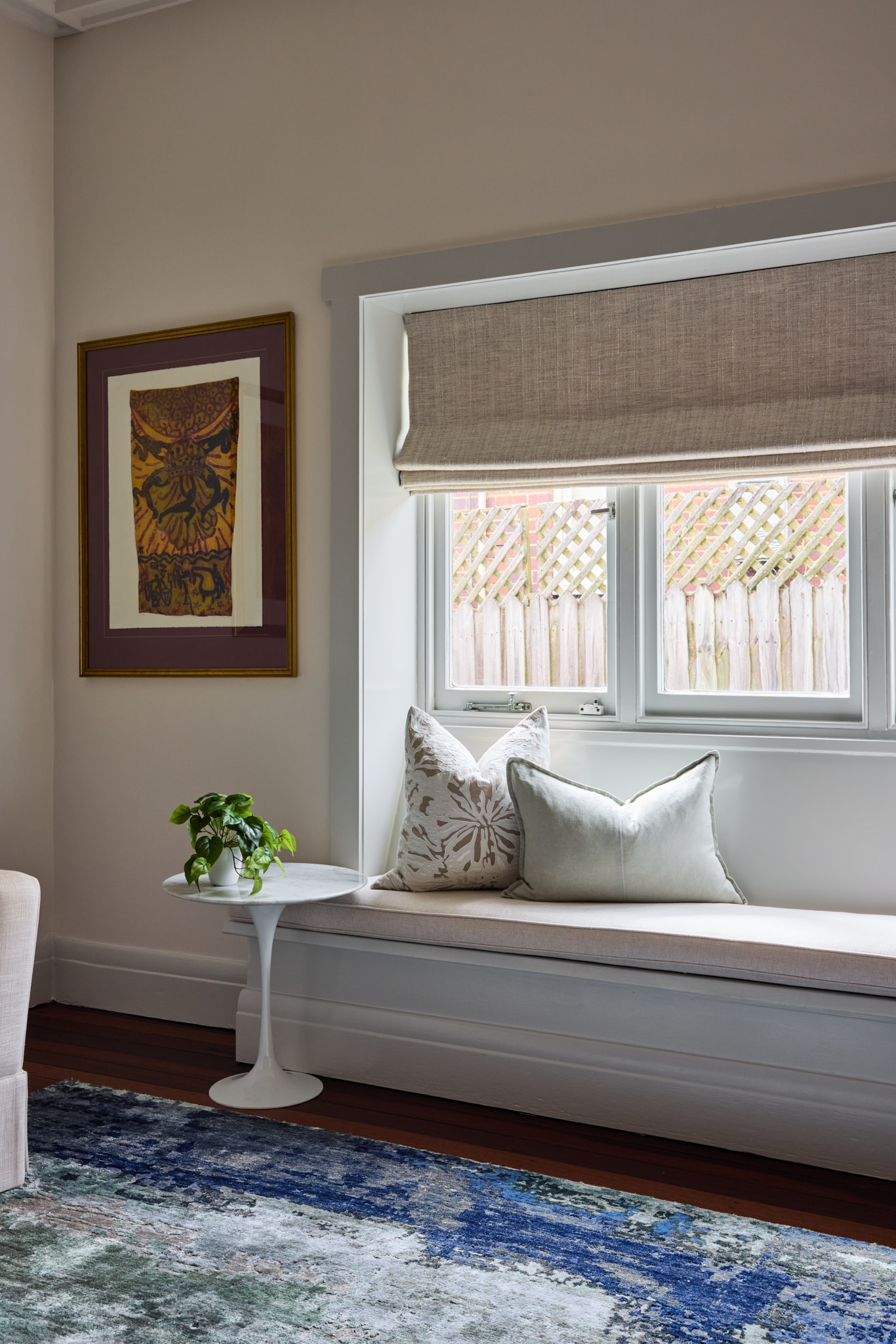 A cozy window seat with neutral cushions and a small plant sits beside a beige-shaded window, while a framed artwork hangs above a blue-patterned rug in this charming Mosman retreat.