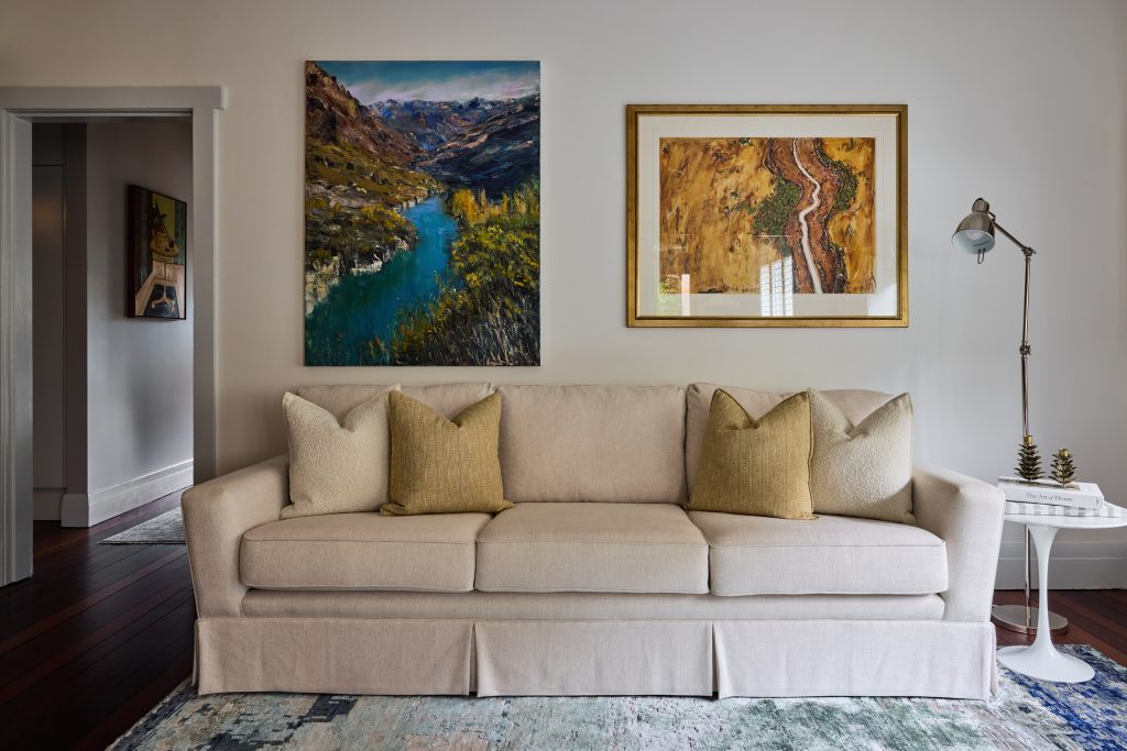 Neutral-toned sofa with four beige pillows against a wall, landscape paintings above, and a floor lamp plus small round table on the right refresh this Mosman home.