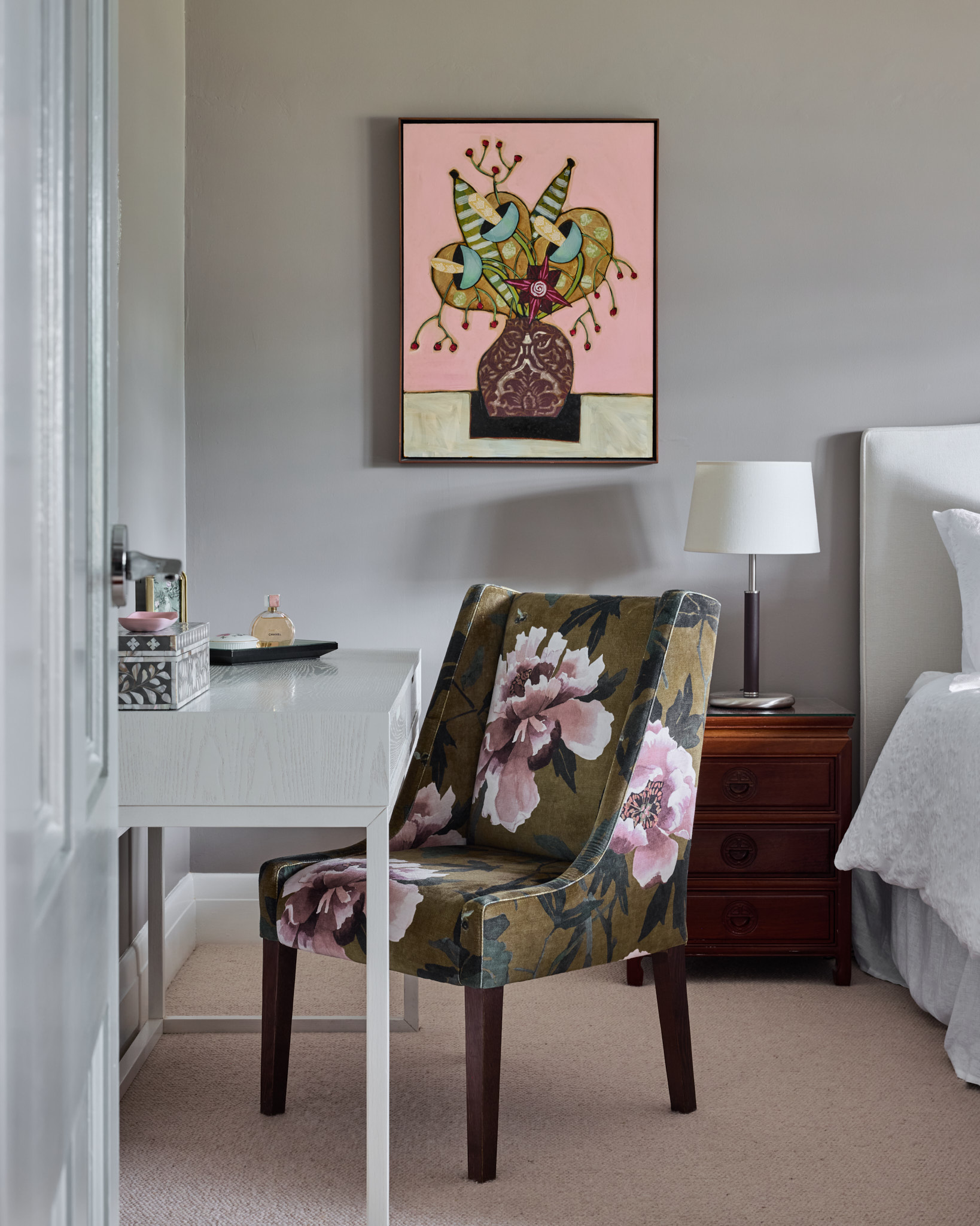 Floral upholstered chair at a white desk next to a wooden nightstand with lamp, below framed abstract flower art on a light gray wall.