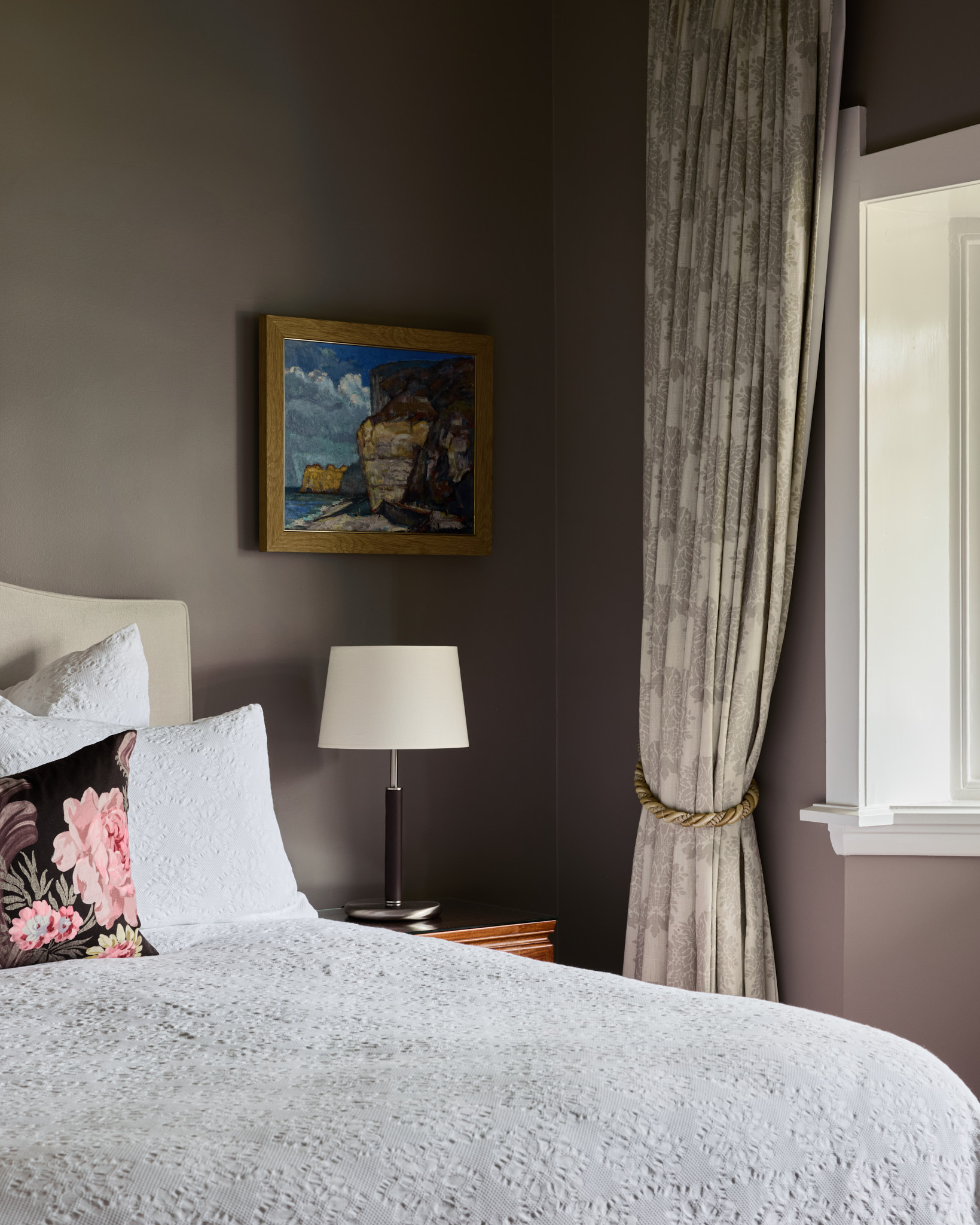 A neatly made bed with white linens, a floral pillow, a side table lamp, a framed painting, and patterned curtains fill this inviting Mosman bedroom.