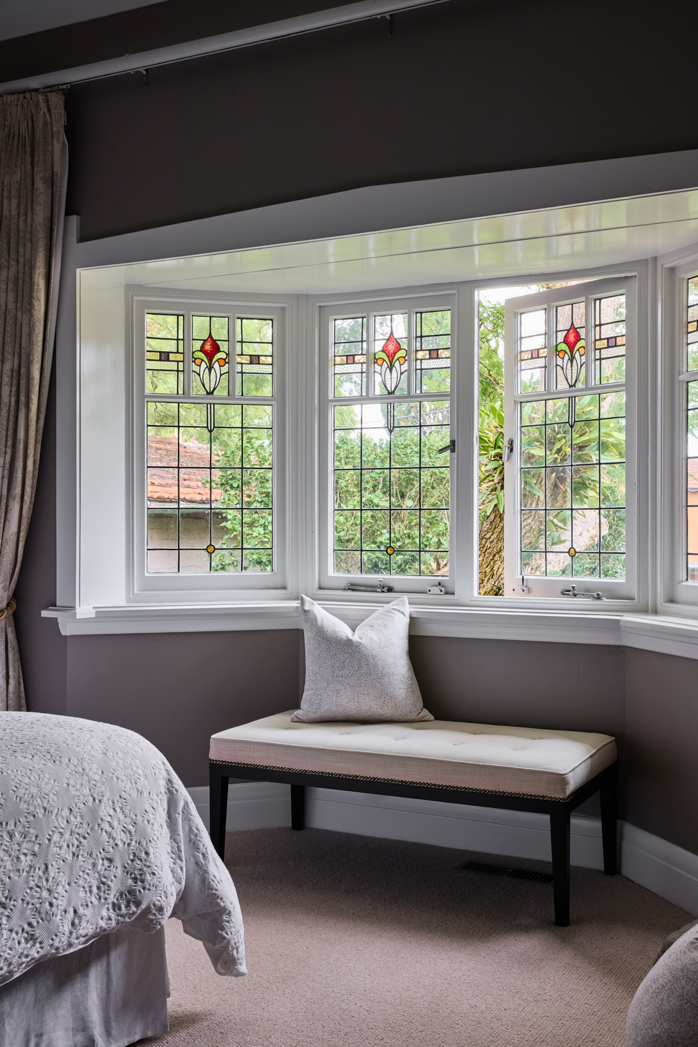 A bench with a white cushion and pillow is positioned below stained glass windows in a carpeted bedroom with gray walls and beige curtains.