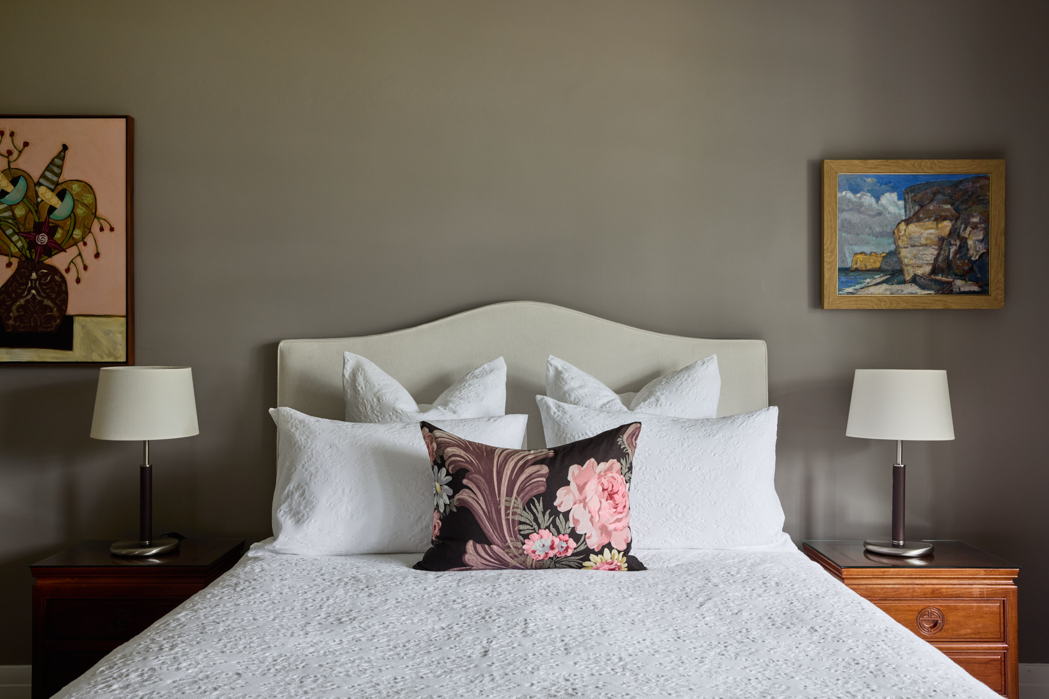 A neatly made bed with white bedding and a floral pillow sits between two wooden nightstands with lamps, set against a gray wall with two paintings.