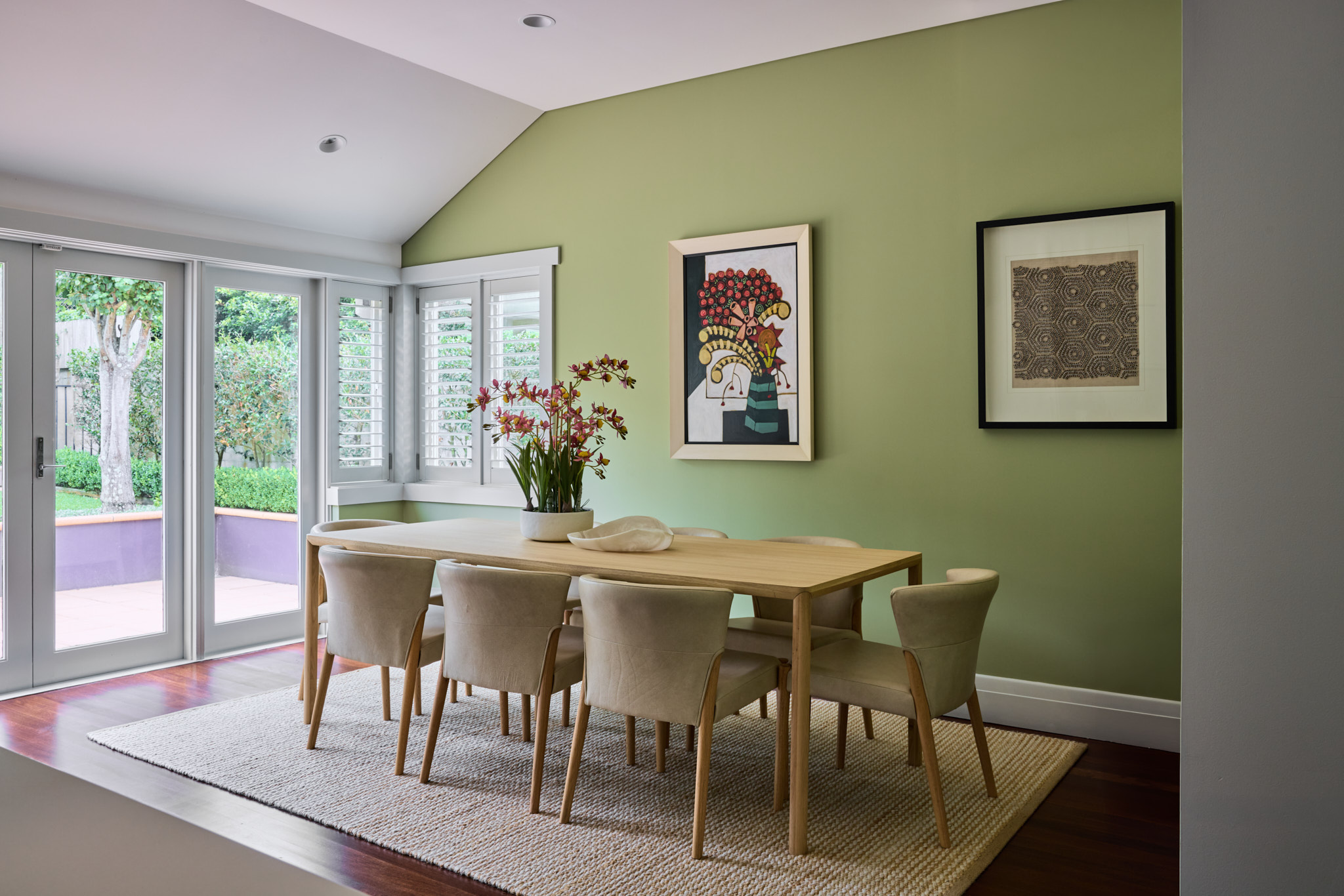 Modern dining room with a light wood table, six beige chairs, green accent wall with two artworks, and large windows overlooking a garden.