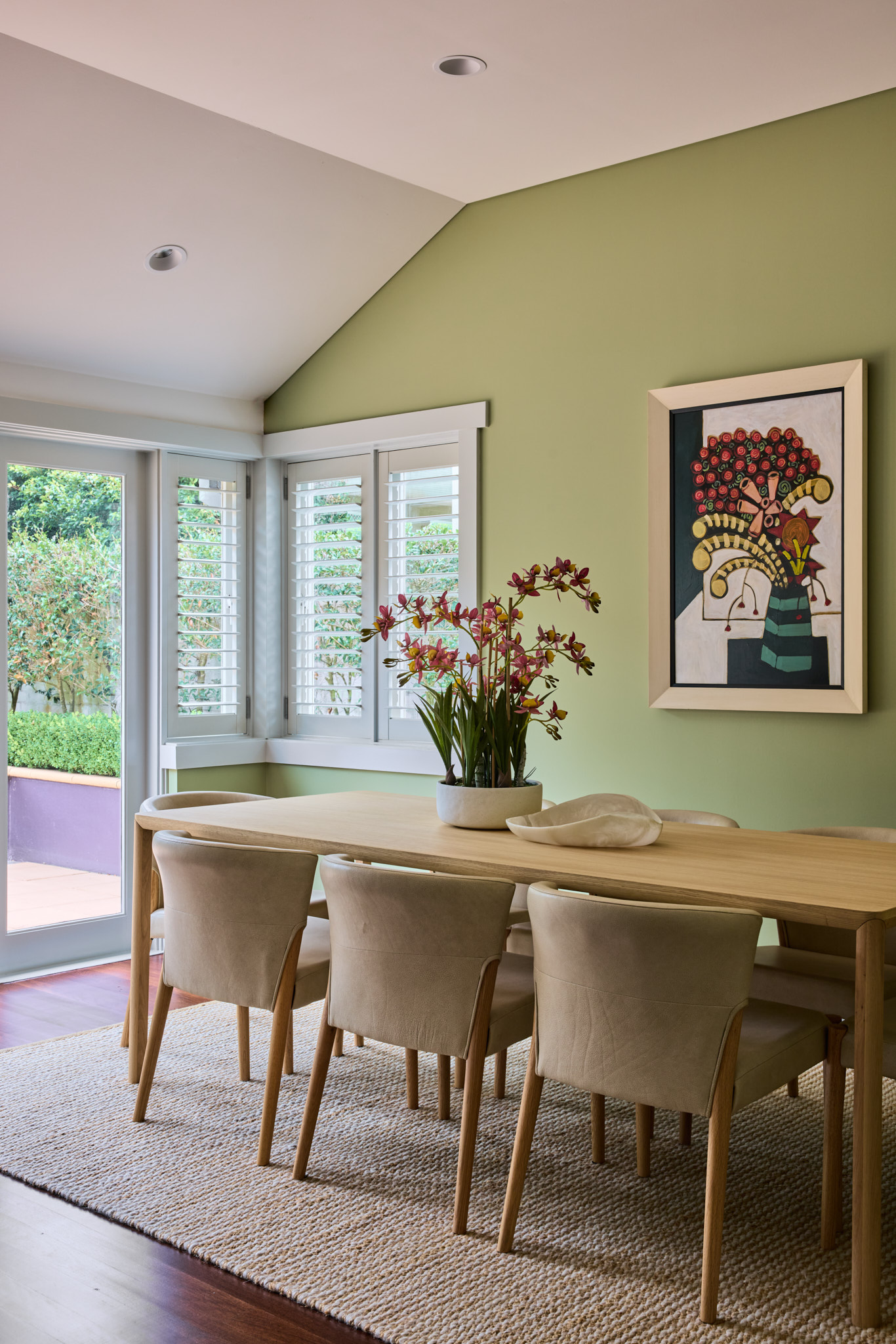 A dining area with a light wood table, six beige chairs, a potted flower centerpiece, green walls, an abstract painting, and large windows with white shutters reflects Mosman-inspired design.