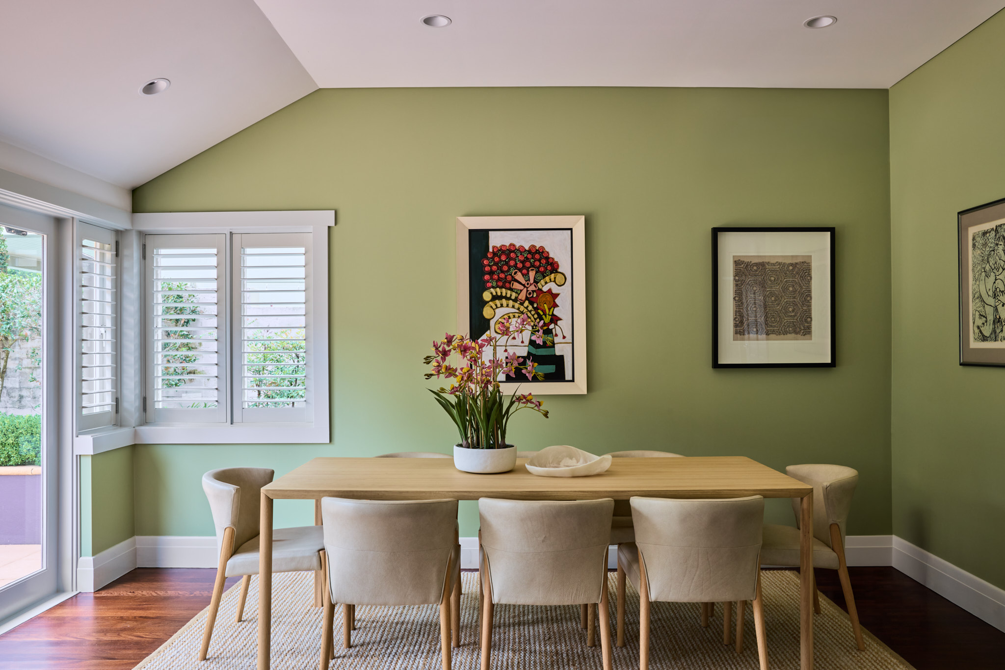 Modern Mosman dining room featuring a light wood table, six cream chairs, green walls, three framed artworks, a window with white shutters, and a potted orchid centerpiece.