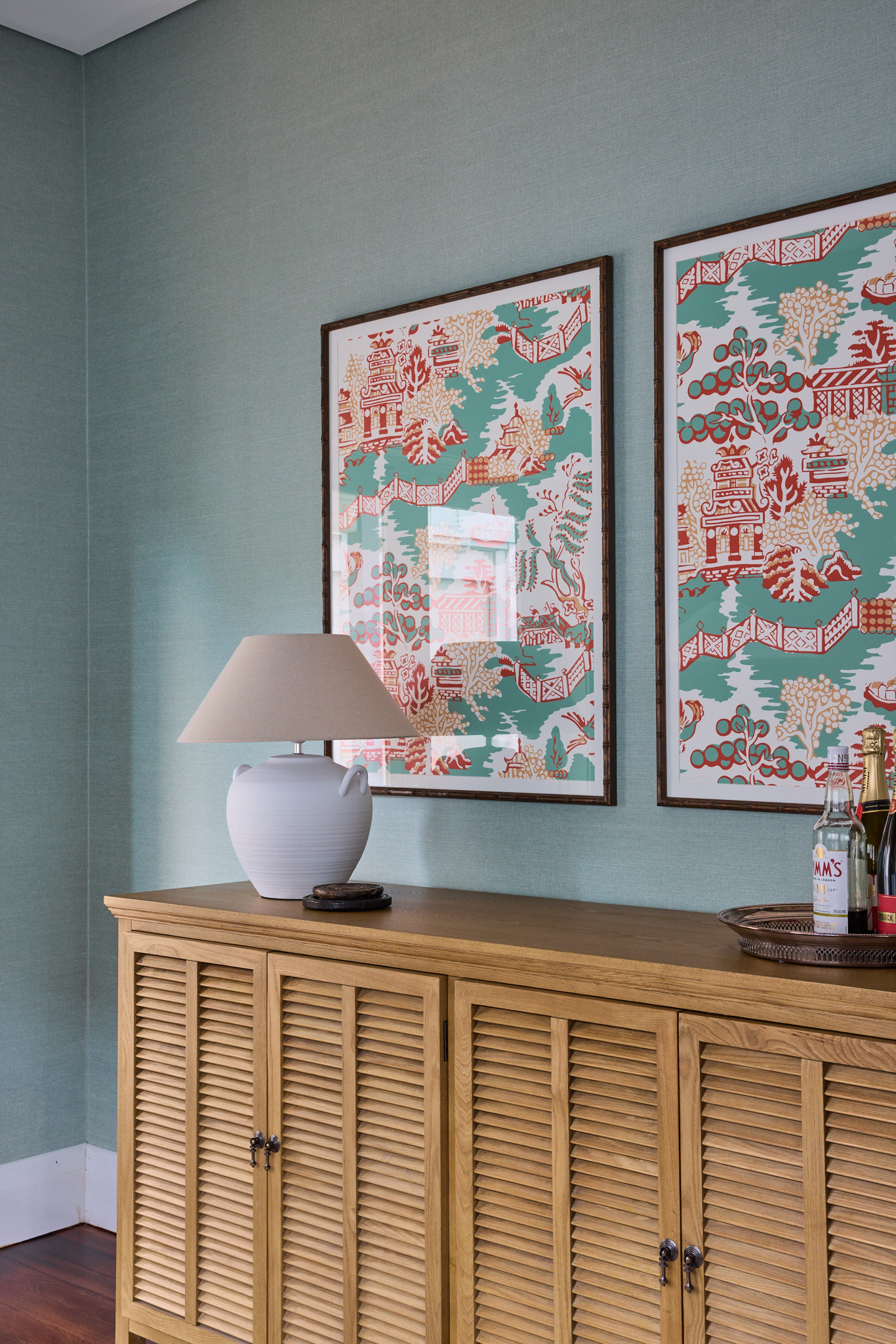A wooden cabinet with a lamp and bottles sits below two colorful prints on a light blue wall, creating an inviting feel in this Mosman home.