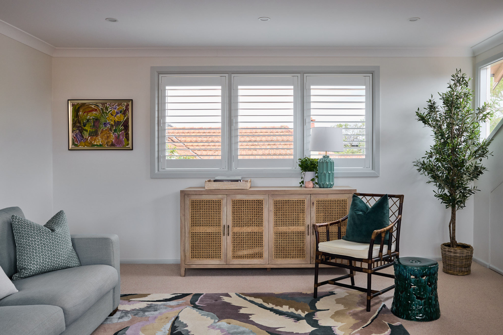Bright living room with grey sofa, patterned rug, wicker cabinet, potted plant, bamboo chair, lamp, artwork and decorative stool under three shuttered windows—stylish inspiration for a Mosman home refresh.