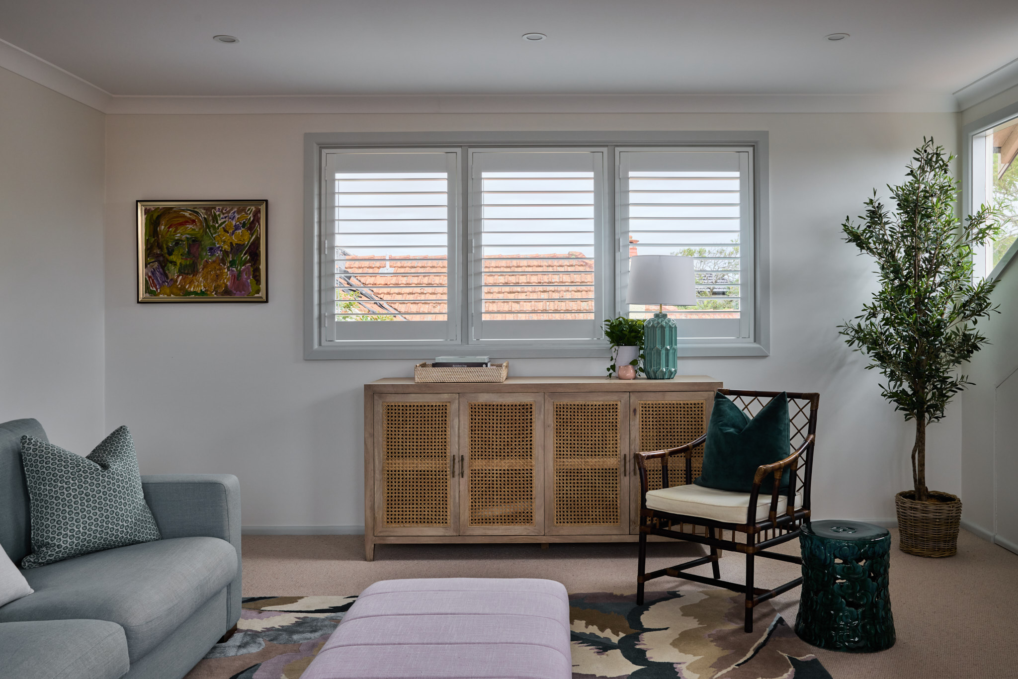 A beautifully renovated Mosman living room features a gray sofa, wooden cabinet, chair, and decor, illuminated by sunlight streaming through three shuttered windows.